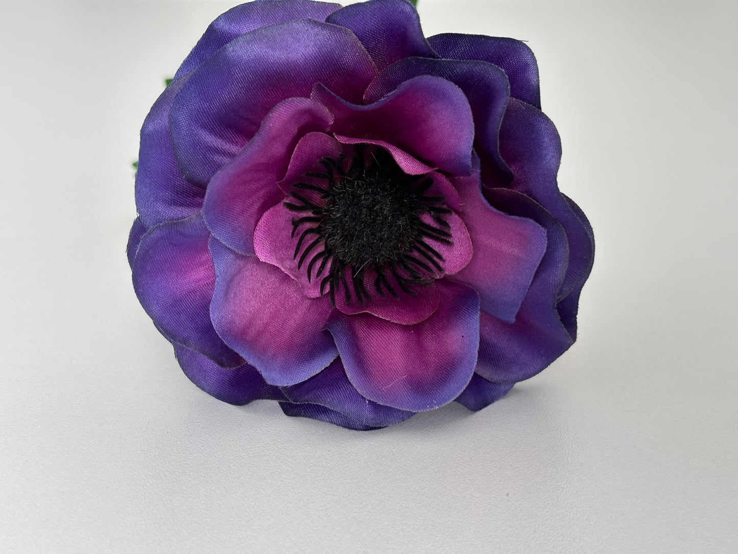 Macro close up overhead shot of an artificial purple anemone flower head showing the stunning layered deep purple and magenta silk petals with a striking dark black spiky centre on a plain white background