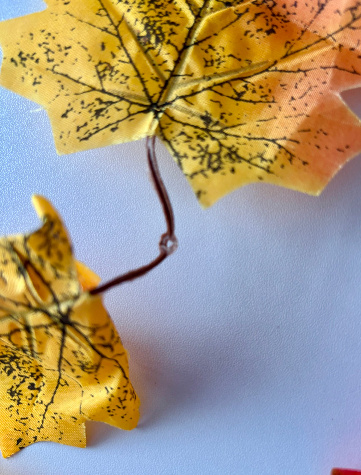 Close up detail shot of a detachable double silk maple leaf section from an autumn maple leaf garland showing rich golden yellow leaves with detailed black leaf vein markings attached to the brown flexible plastic garland strand against a plain pale blue background
