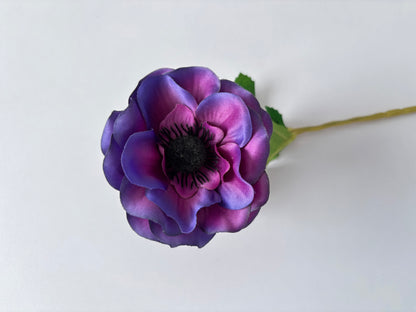Close up landscape shot of an artificial purple anemone flower head showing the beautiful ruffled layered deep purple and magenta silk petals with a striking dark black spiky centre and green foliage leaf visible behind on a plain white background