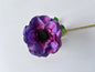 Close up landscape shot of an artificial purple anemone flower head showing the beautiful ruffled layered deep purple and magenta silk petals with a striking dark black spiky centre and green foliage leaf visible behind on a plain white background