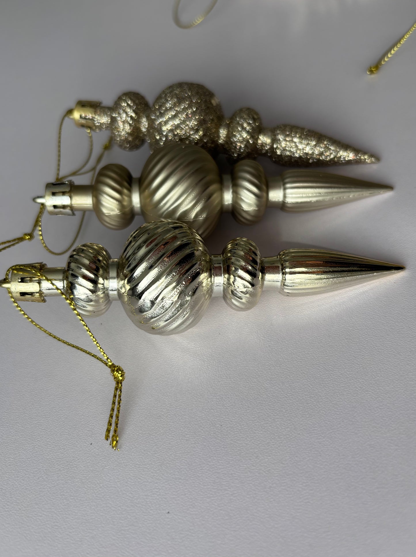 Three champagne gold Christmas spindle baubles arranged diagonally showing metallic and glittered finishes with ribbed bulbous spherical tops, twisted ribbed middles and elongated pointed drop bases hanging from gold twisted cord strings on a plain light grey background