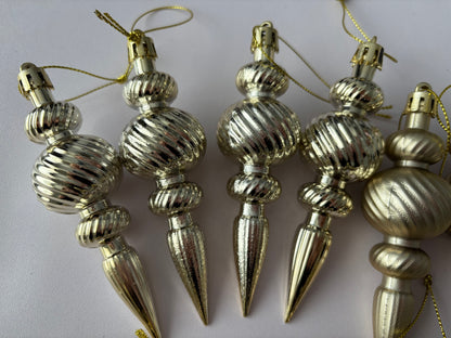 Close up of five champagne gold metallic finish Christmas spindle baubles with highly polished ribbed bulbous spherical tops, twisted ribbed middles and elongated pointed drop bases with gold hanging strings arranged upright on a plain light background