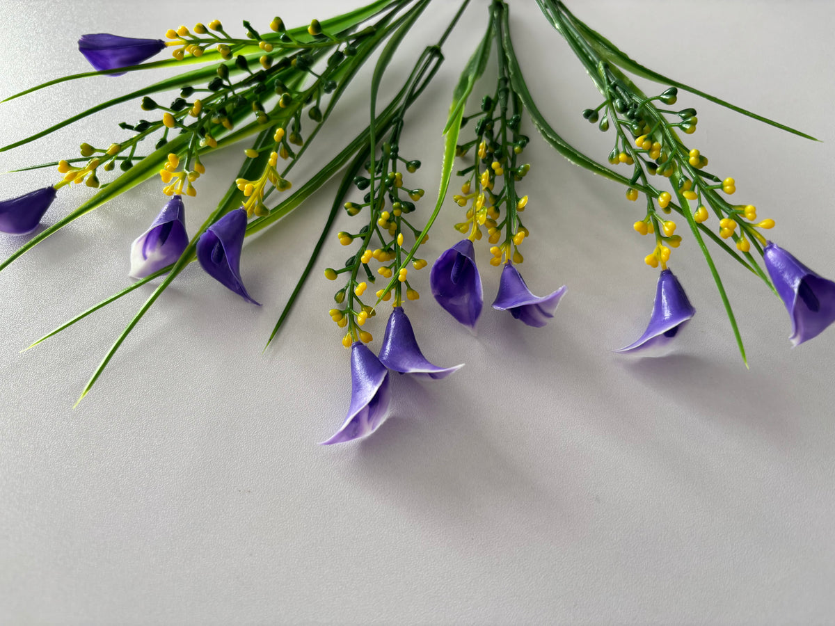 Overhead flat lay shot of an artificial purple small calla lily foliage bunch laid horizontally showing all five individual wired stems spread out with small purple calla lily flower heads drooping naturally downward, clusters of bright yellow berry picks and tall thin green grass stems fanning across against a plain light grey background