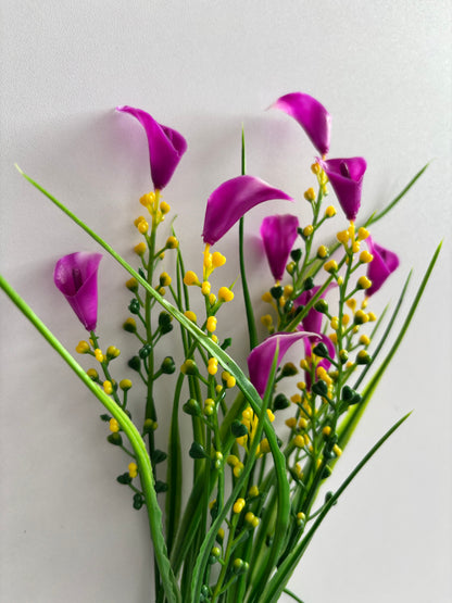 Close up upright shot of an artificial pink small calla lily foliage bunch showing multiple small bright magenta pink calla lily flower heads with white inner petal detail up close alongside dense clusters of small bright yellow berry pick stems and tall thin green grass stems against a plain light grey background