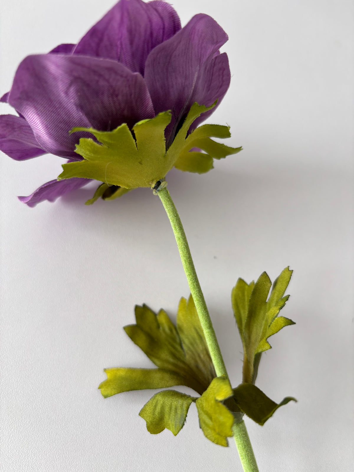 Full length upright portrait shot of an artificial purple silk anemone single stem showing the open purple flower head at the top with bright green deeply serrated detachable foliage leaves attached just below the flower head and a second set of detachable foliage leaves positioned lower down the fabric wrapped green stem against a plain light background