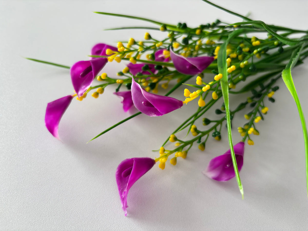 Close up diagonal flat lay shot of an artificial pink small calla lily foliage bunch showing multiple detached small bright magenta pink calla lily flower heads with white inner petal detail scattered across the foreground with bright yellow berry pick stems and tall green grass stems visible behind against a plain white surface