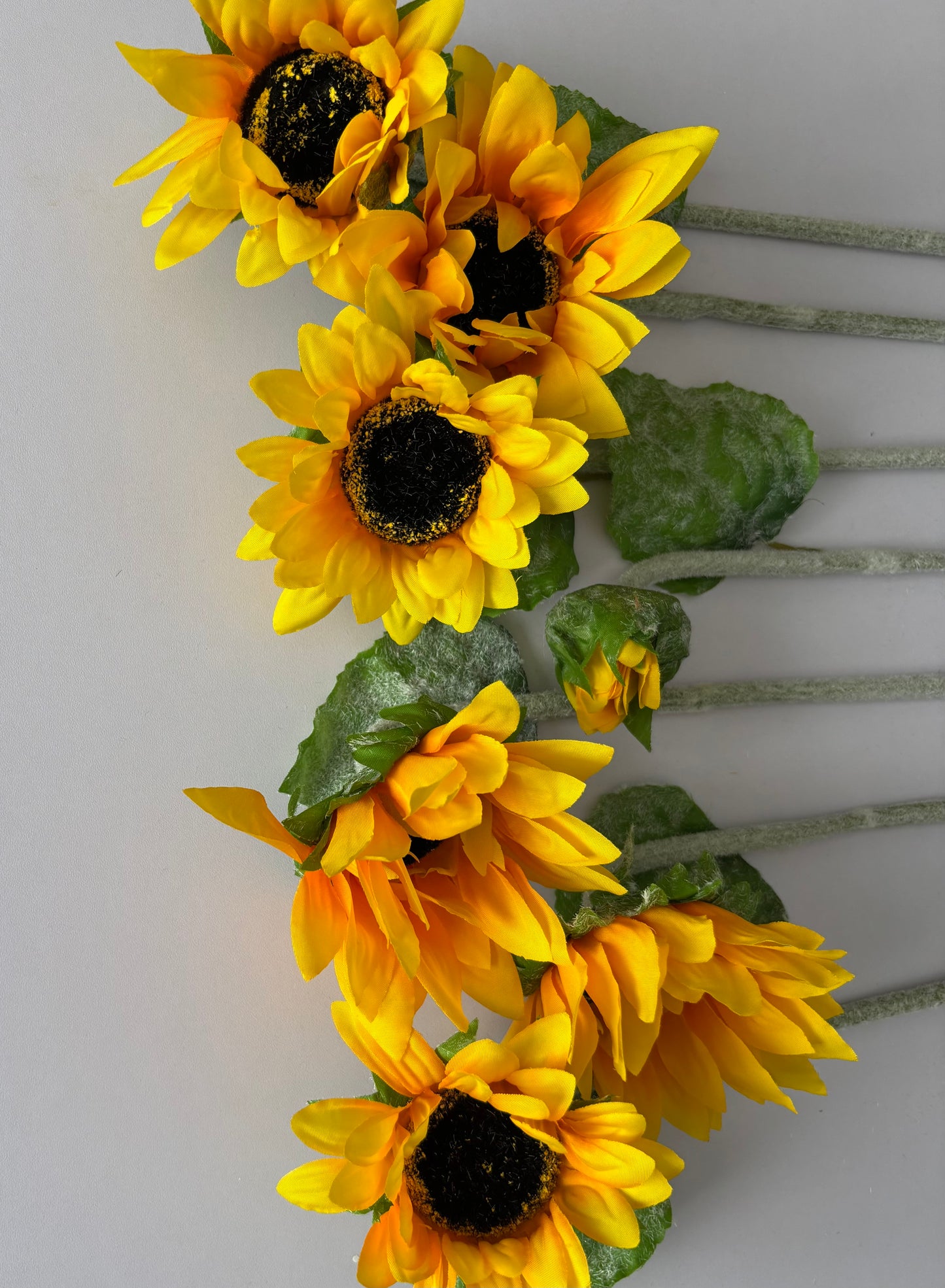 Seven artificial sunflower stems arranged diagonally showing the mixed size golden yellow flower heads with dark brown centres and lush green foliage cascading downwards with glittery silver textured stems on a plain light grey background