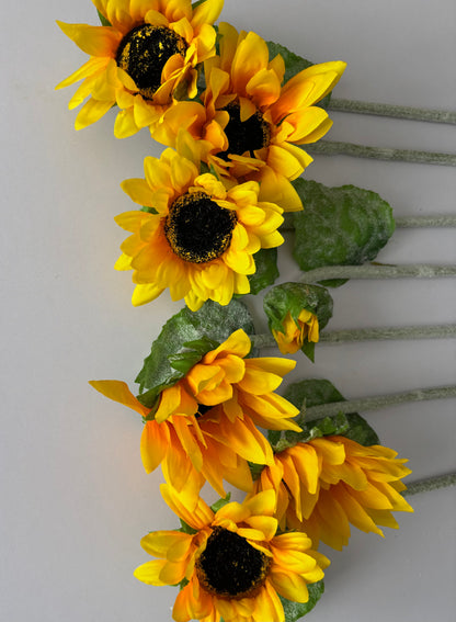 Seven artificial sunflower stems arranged diagonally showing the mixed size golden yellow flower heads with dark brown centres and lush green foliage cascading downwards with glittery silver textured stems on a plain light grey background