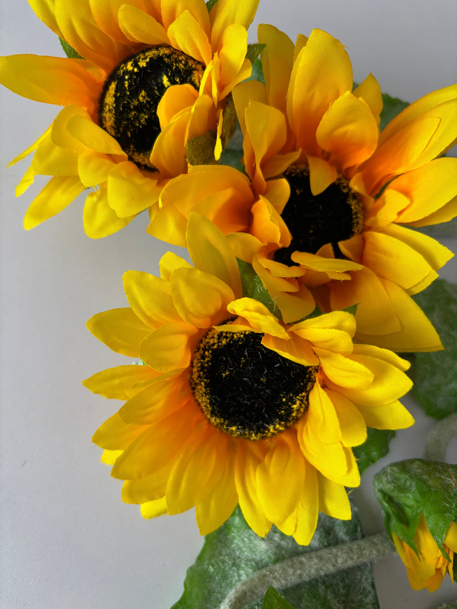 Macro close up shot of two artificial sunflower heads showing the intricate golden yellow petals with orange tones and dark black textured centre detail with bright green foliage on a plain light grey background