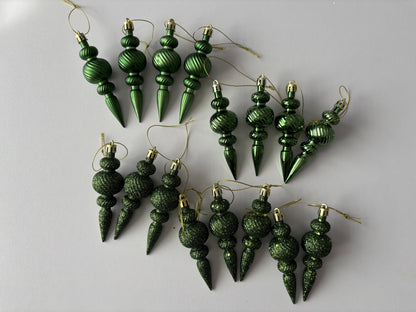 Flat lay of all 16 green Christmas spindle baubles in matte, metallic and glittered finishes with ribbed spherical tops, beaded middles and elongated pointed drop bases with gold hanging strings arranged on a plain light grey background