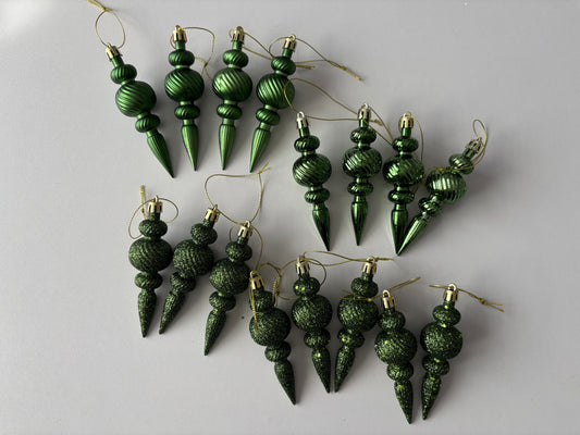 Flat lay of all 16 green Christmas spindle baubles in matte, metallic and glittered finishes with ribbed spherical tops, beaded middles and elongated pointed drop bases with gold hanging strings arranged on a plain light grey background
