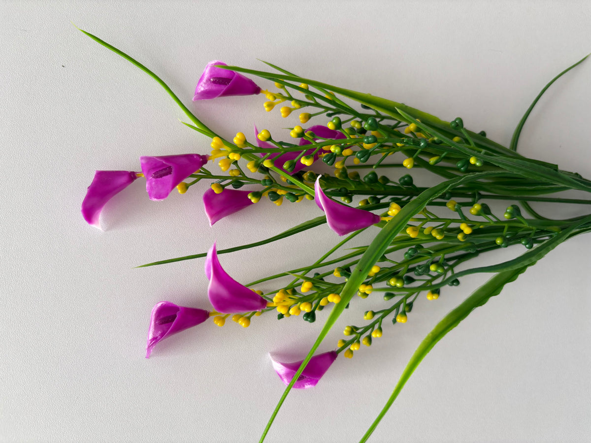 Diagonal flat lay shot of an artificial pink small calla lily foliage bunch laid across a plain white surface from a slightly different angle showing multiple detached small bright magenta pink calla lily flower heads scattered across the foreground with bright yellow berry pick clusters and tall thin green grass stems fanning outward above