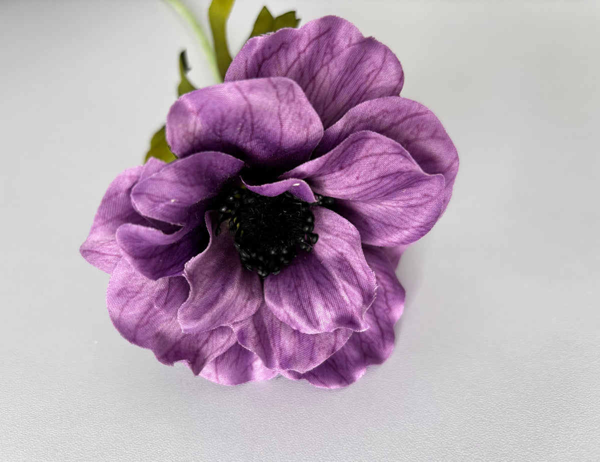 Close up front facing shot of a large artificial purple silk anemone flower head showing beautifully open layered cup-shaped petals with exquisite vein detail throughout the silk and a dramatic raised dark black seed centre with tiny black stamens against a plain light grey background with bright green foliage visible behind