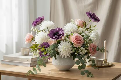 AI lifestyle image of a lush mixed spring floral table centrepiece arrangement in a white ceramic bowl vase featuring purple anemones, large white chrysanthemums, blush pink roses and trailing green eucalyptus displayed on a light wooden table with a white candle and stacked books in a softly lit room