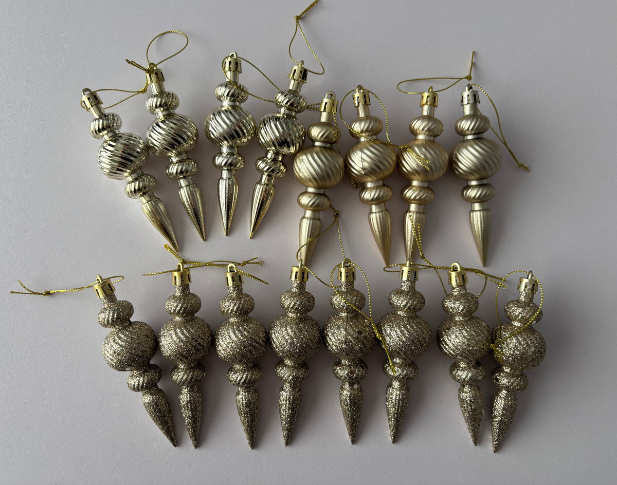 Flat lay of all 16 champagne gold Christmas spindle baubles in matte and glittered finishes arranged in two rows with ribbed spherical tops, twisted middles and elongated pointed drop bases with gold hanging strings on a plain light grey background