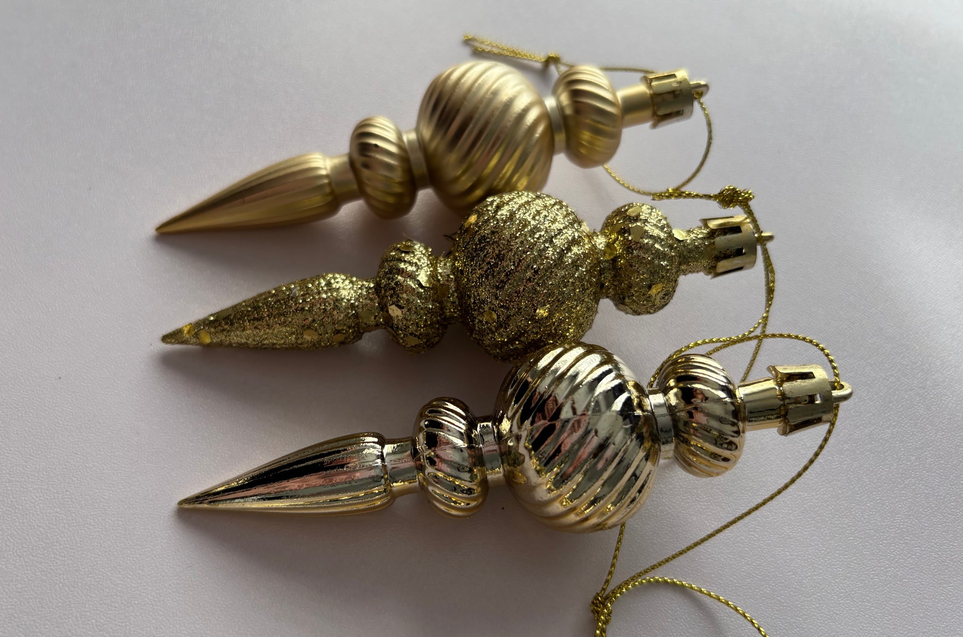 Three gold Christmas spindle baubles stacked vertically showing matte, glittered and metallic finishes with ribbed spherical tops, beaded middles and elongated pointed drop bases with gold hanging hooks on a plain light grey background