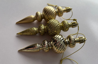 Three gold Christmas spindle baubles stacked vertically showing matte, glittered and metallic finishes with ribbed spherical tops, beaded middles and elongated pointed drop bases with gold hanging hooks on a plain light grey background