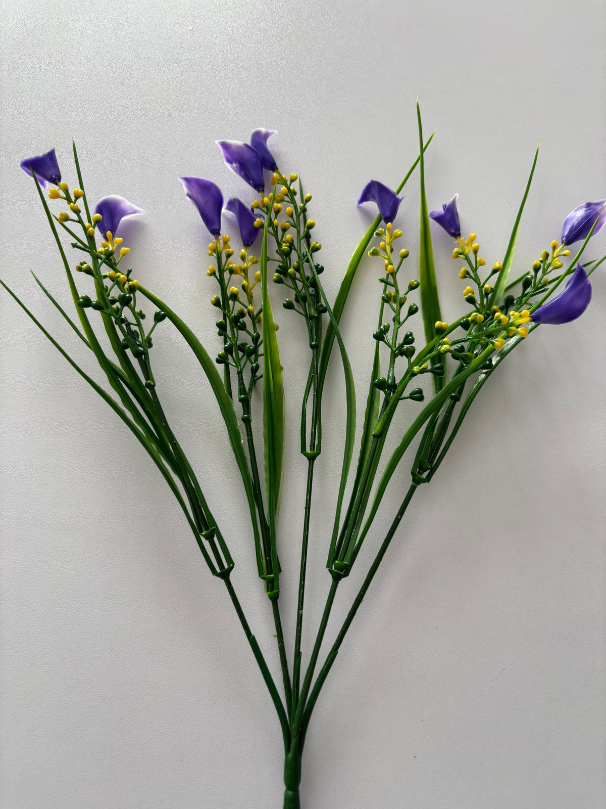 Upright portrait shot of an artificial purple small calla lily foliage bunch showing the full bunch from base to tip with the single bottom stem clearly visible at the base and five individual wired stems fanning out above with small purple calla lily flower heads, bright yellow berry pick stems and tall thin green grass stems against a plain light background