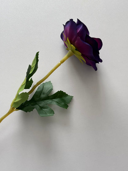 Overhead flat lay shot of an artificial purple anemone single stem laid diagonally showing the closed partially open deep purple and violet flower bud at the top with yellow-green wired stem and lush dark green serrated foliage leaves fanned out at the bottom on a plain light grey background