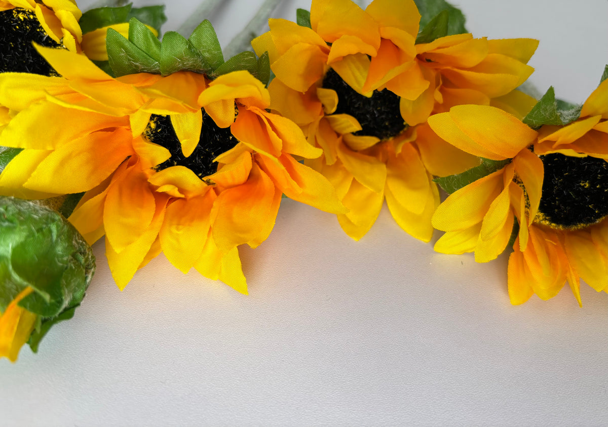 Overhead close up shot of multiple artificial sunflower heads showing the large golden yellow and deep orange petals with dark brown to black textured centres and lush bright green foliage on a plain white background
