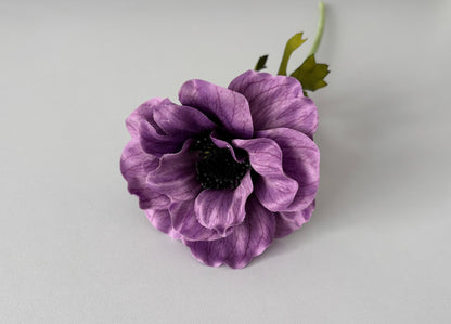 Overhead angled shot of an artificial purple silk anemone flower head showing beautifully open layered cup-shaped petals with fine vein detail throughout, a dramatic dark black raised centre and bright green detachable serrated foliage leaves visible behind against a plain light grey background