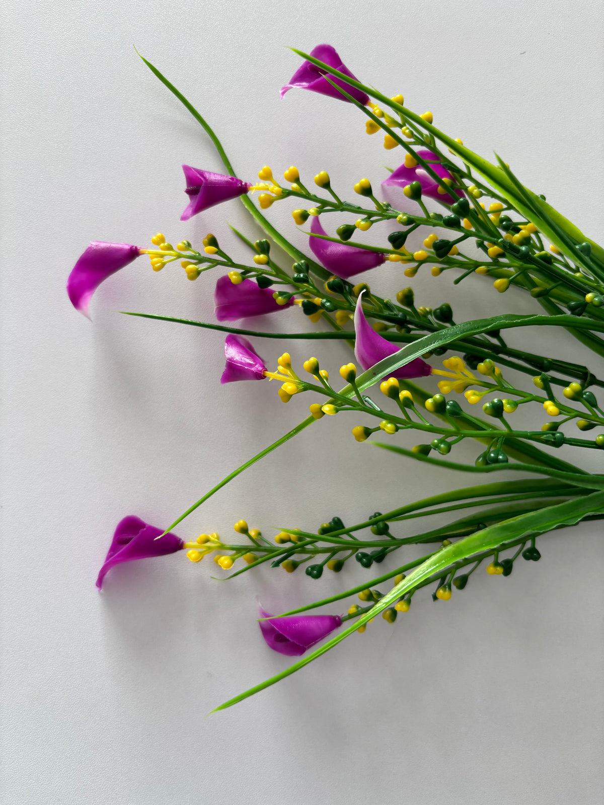 Angled upright shot of an artificial pink small calla lily foliage bunch from a slightly overhead perspective showing the full bunch spread with small bright magenta pink calla lily flower heads dotted throughout dense clusters of bright yellow berry pick stems and tall thin green grass stems all fanning out from a single bottom stem against a plain light grey background