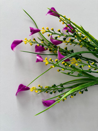 Angled upright shot of an artificial pink small calla lily foliage bunch from a slightly overhead perspective showing the full bunch spread with small bright magenta pink calla lily flower heads dotted throughout dense clusters of bright yellow berry pick stems and tall thin green grass stems all fanning out from a single bottom stem against a plain light grey background