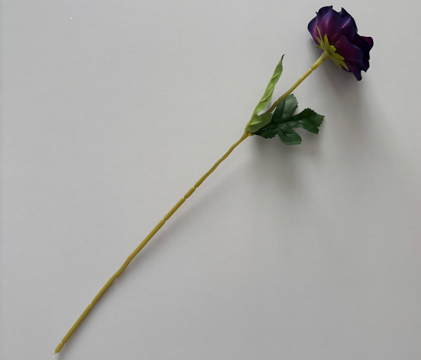 Overhead flat lay shot of an artificial purple anemone single stem laid diagonally across the full frame showing the complete stem length with closed deep purple and violet flower bud at the top right dark green serrated foliage leaves in the middle and the full length yellow-green wired stem extending to the bottom left on a plain light grey background