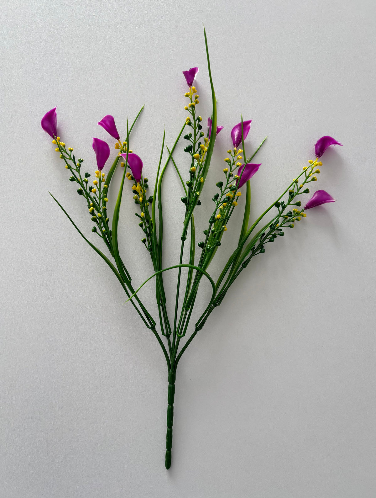 Upright full spread shot of an artificial pink small calla lily foliage bunch showing five individual wired stems fanning out from a single bottom stem with small bright magenta pink calla lily flower heads, tall thin green grass stems and small bright yellow berry pick stems all arranged against a plain light grey background