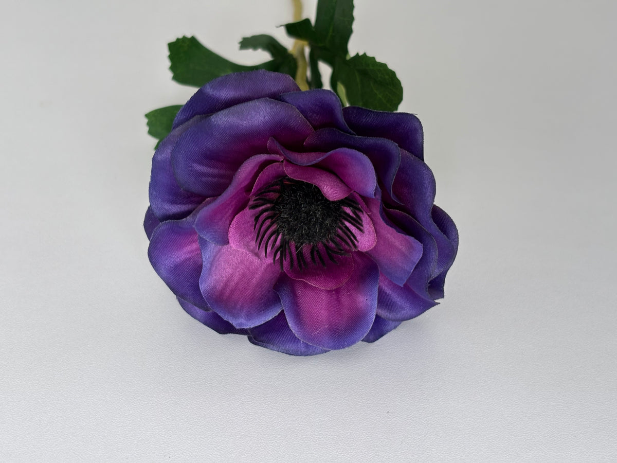 Close up front facing shot of an artificial purple anemone flower head showing the stunning layered deep purple and magenta ruffled silk petals with a dramatic dark black spiky centre and dark green serrated foliage leaves visible behind on a plain light background