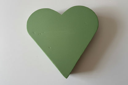 Overhead flat lay shot of a green heart shaped oasis wet floral foam frame showing the full heart shape and solid two-layer foam construction with soft insertable wet foam top surface against a plain white background