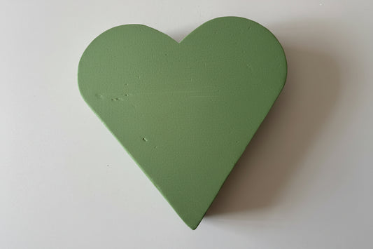 Overhead flat lay shot of a green heart shaped oasis wet floral foam frame showing the full heart shape and solid two-layer foam construction with soft insertable wet foam top surface against a plain white background