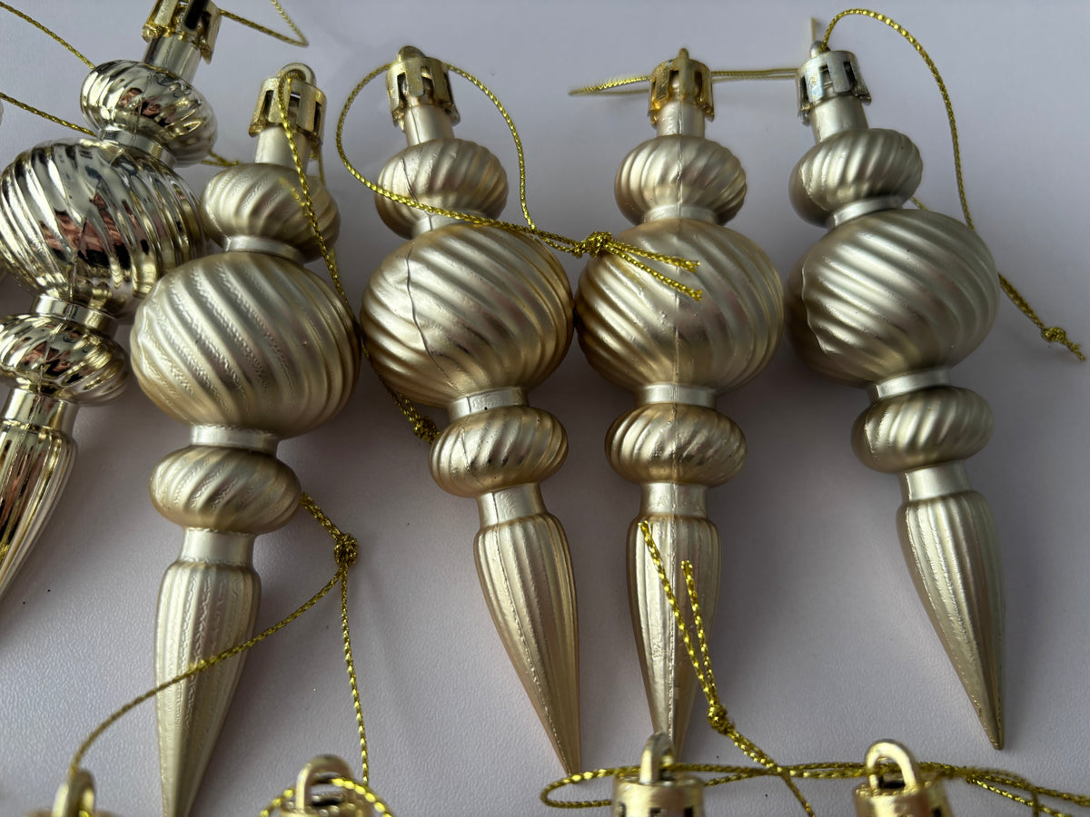 Close up of five champagne gold matte finish Christmas spindle baubles with ribbed bulbous spherical tops, twisted ribbed middles and elongated pointed drop bases with gold hanging strings laid horizontally on a plain light background