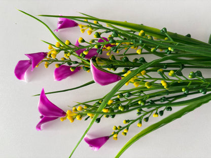 Diagonal flat lay shot of an artificial pink small calla lily foliage bunch laid across a plain white surface showing the full stem length with multiple detached small bright magenta pink calla lily flower heads scattered in the foreground and bright yellow berry pick stems and tall thin green grass stems spreading outward from the single bottom stem