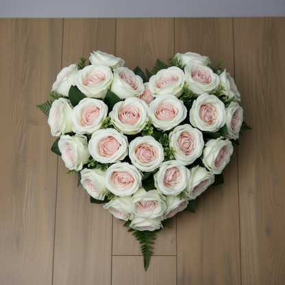 Artificial cream and pink cabbage roses styled in a heart shaped funeral tribute with white and blush pink roses and green fern foliage on a light wood floor