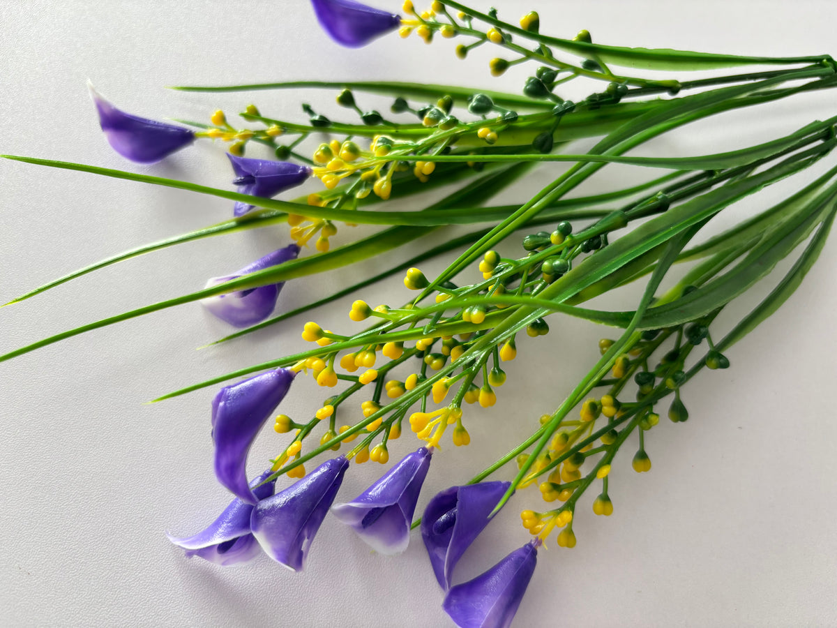 Close up diagonal flat lay shot of an artificial purple small calla lily foliage bunch showing multiple detached small purple calla lily flower heads with violet and white inner petal detail scattered in the foreground with bright yellow berry pick clusters and tall green grass stems spreading across a plain white background
