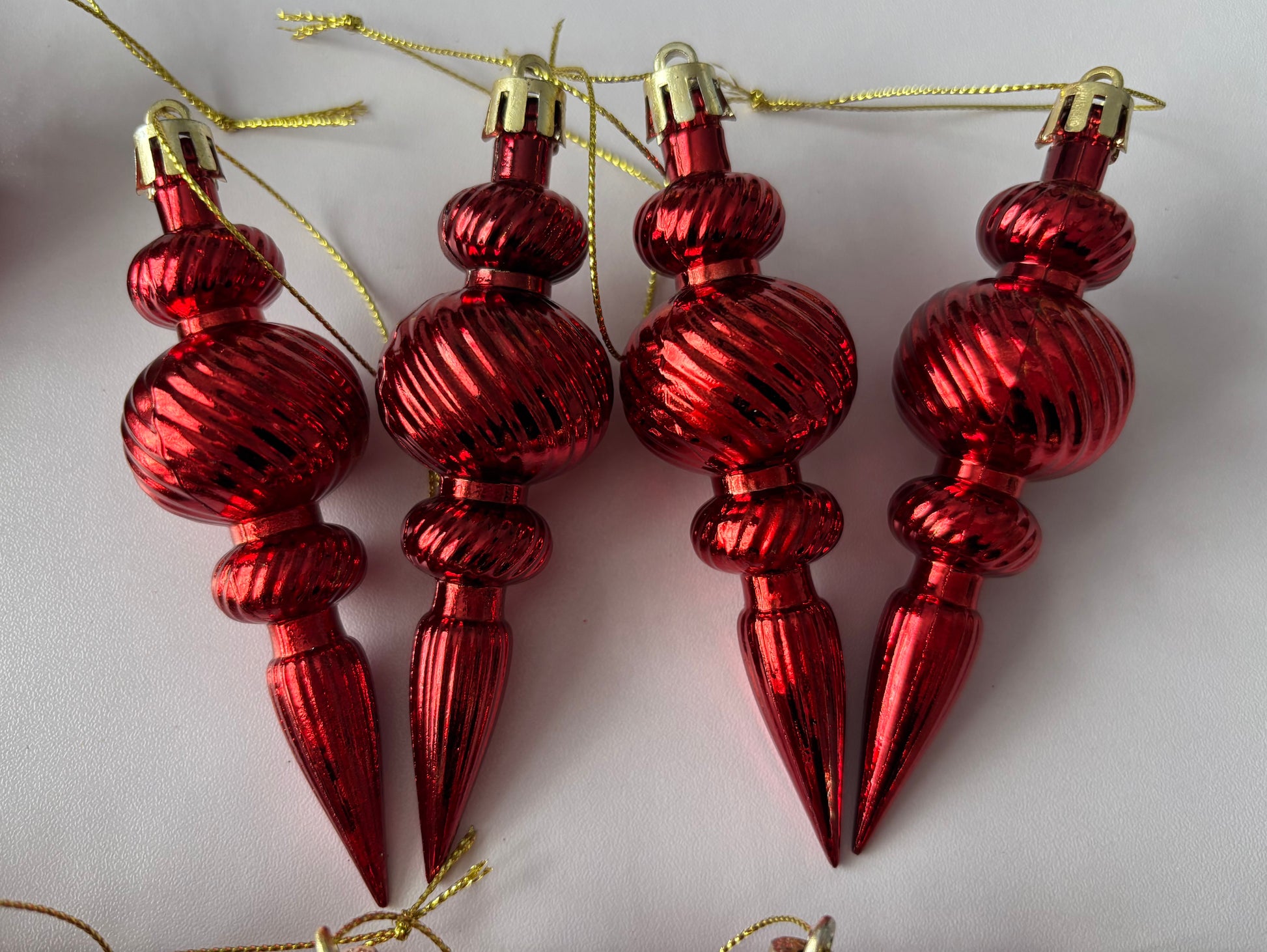 Close up of four bright red metallic Christmas spindle baubles with highly polished ribbed spherical tops, ribbed middles and elongated pointed drop bases with gold hanging strings arranged on a plain light background