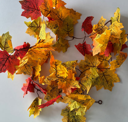 Overhead flat lay shot of an autumn maple leaf garland 180cm with multiple detachable double silk maple leaf sections in rich yellow, orange, red and green tones spread across a plain light grey background showing the full garland arrangement with brown plastic garland strand and hanging hoop visible at the bottom right