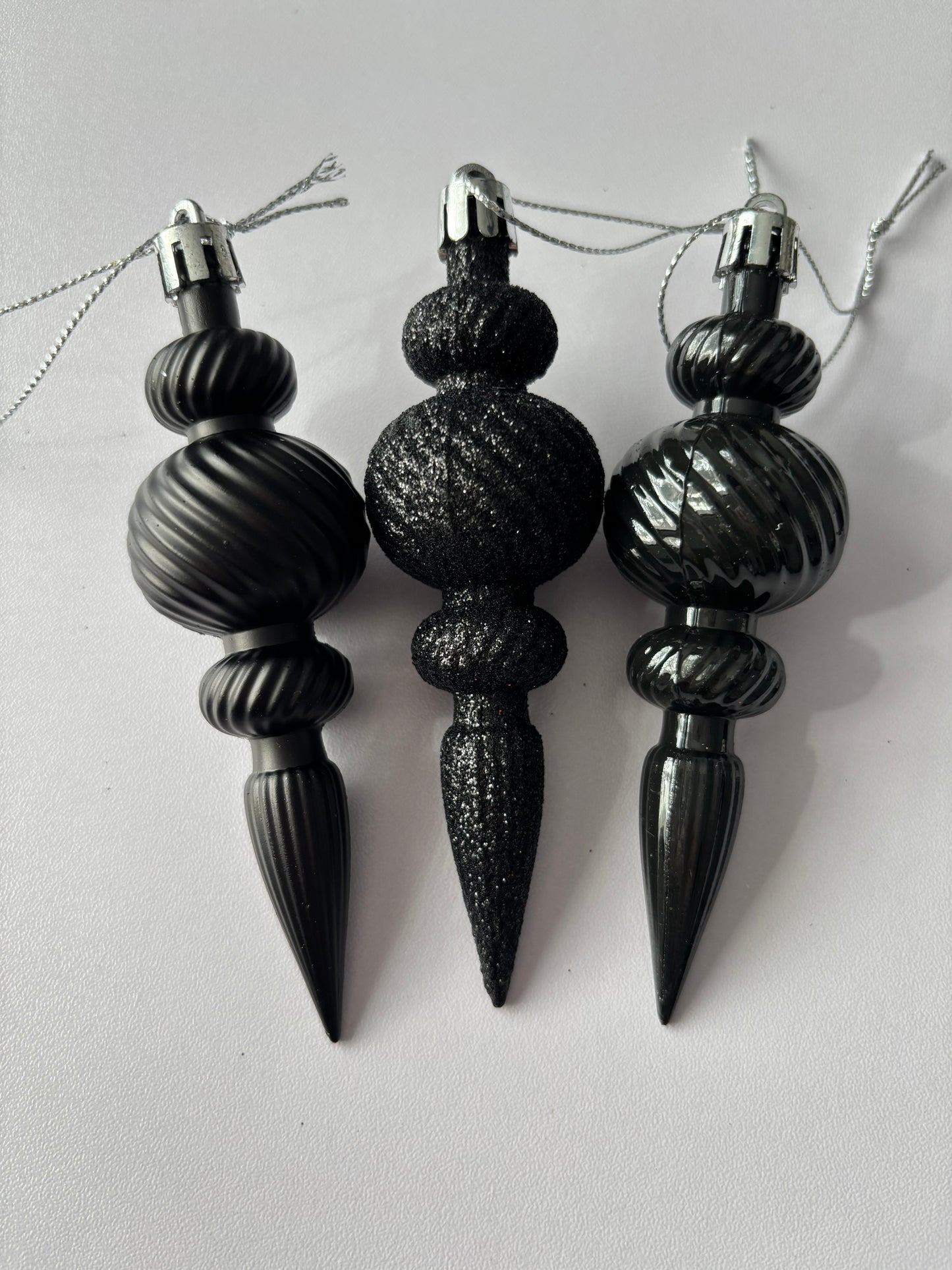 Three black Christmas icicle finial baubles standing upright showing matte, glittered and metallic finishes with ribbed spherical tops, beaded middles and elongated pointed drop bases with silver hanging loops on a plain white background