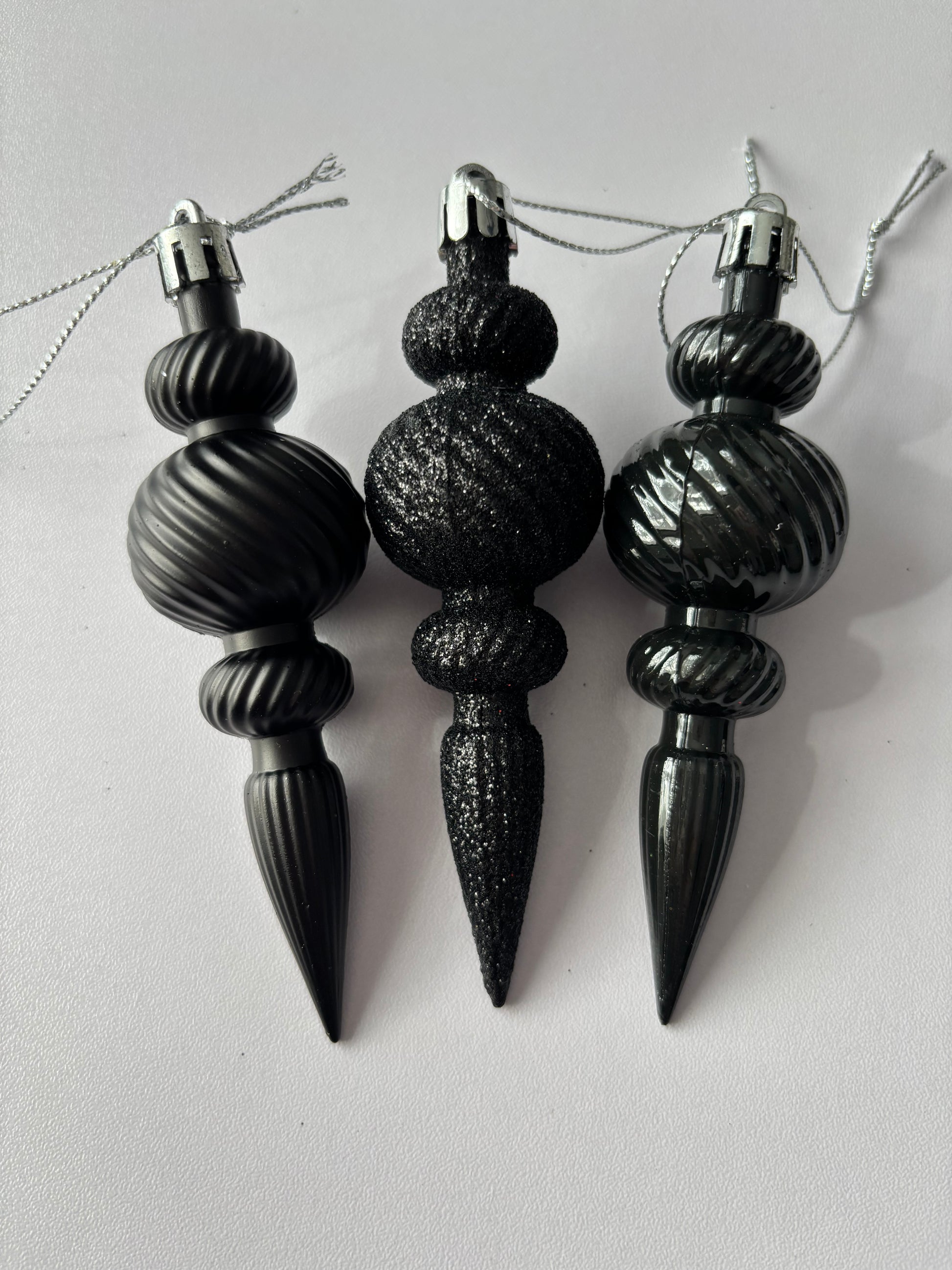 Three black Christmas icicle finial baubles standing upright showing matte, glittered and metallic finishes with ribbed spherical tops, beaded middles and elongated pointed drop bases with silver hanging loops on a plain white background