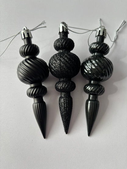 Three black Christmas icicle finial baubles standing upright showing matte, glittered and metallic finishes with ribbed spherical tops, beaded middles and elongated pointed drop bases with silver hanging loops on a plain white background