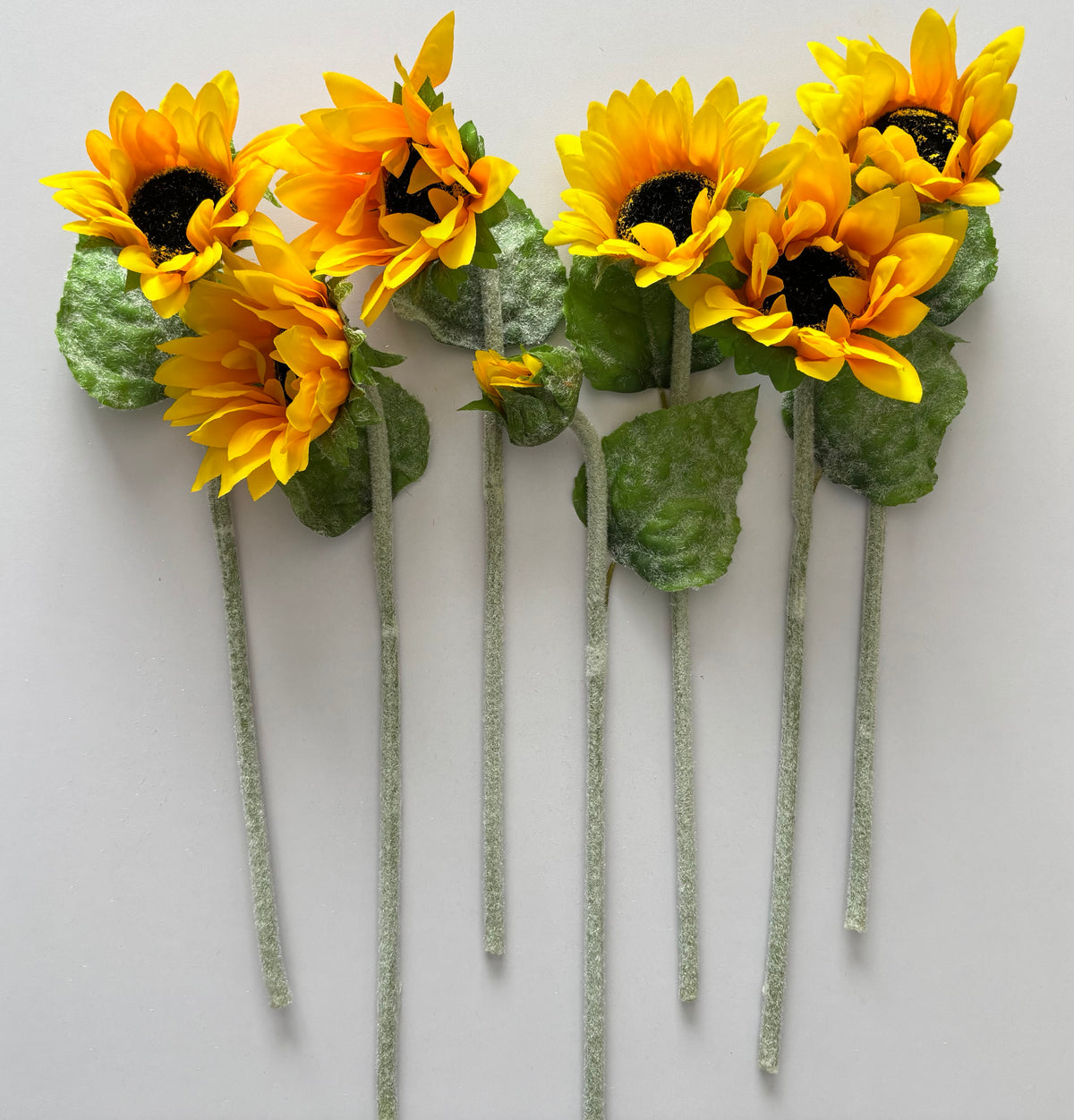 Seven artificial sunflower stems laid flat in a fan arrangement showing the full golden yellow petals dark brown centres lush green foliage and glittery silver textured stems on a plain white background