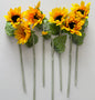 Seven artificial sunflower stems laid flat in a fan arrangement showing the full golden yellow petals dark brown centres lush green foliage and glittery silver textured stems on a plain white background