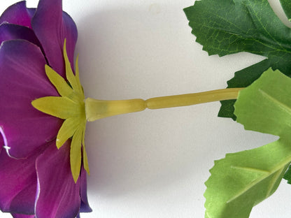 Extreme macro close up underside shot of an artificial purple anemone flower head showing the bright yellow star-shaped plastic calyx and yellow-green wired stem attachment point with deep purple petals and lush dark green serrated foliage leaves on a plain white background