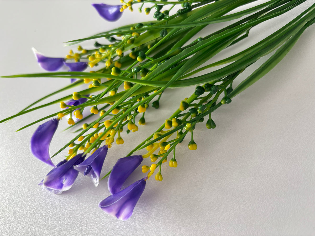 Diagonal flat lay shot of an artificial purple small calla lily foliage bunch laid across a plain light background from a wider angle showing multiple detached small purple calla lily flower heads scattered in the foreground with bright yellow berry pick clusters and tall thin green grass stems spreading outward above