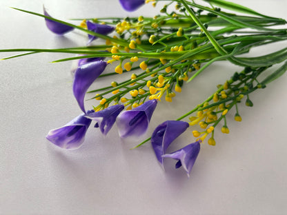 Close up diagonal flat lay shot of an artificial purple small calla lily foliage bunch laid horizontally showing multiple detached small purple calla lily flower heads with beautiful violet and white inner petal detail scattered in front of the bunch with bright yellow berry pick stems and tall green grass stems visible behind against a plain white surface