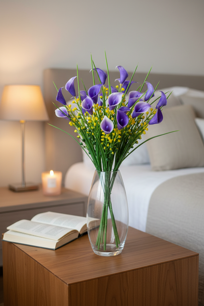 AI lifestyle image of an artificial purple small calla lily foliage bunch displayed upright in a clear glass vase on a warm wooden bedside table with a softly glowing candle and open book in a warmly lit bedroom interior showing the full bunch height with purple calla lily heads, yellow berry picks and tall green grass stems