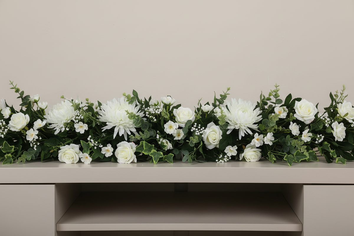 Artificial white chrysanthemum single stems styled in a long lush wedding console table garland with white roses, gypsophila and trailing green eucalyptus foliage arranged across a light beige console table against a plain beige wall