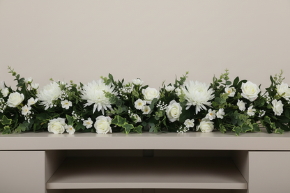 Artificial white chrysanthemum single stems styled in a long lush wedding console table garland with white roses, gypsophila and trailing green eucalyptus foliage arranged across a light beige console table against a plain beige wall