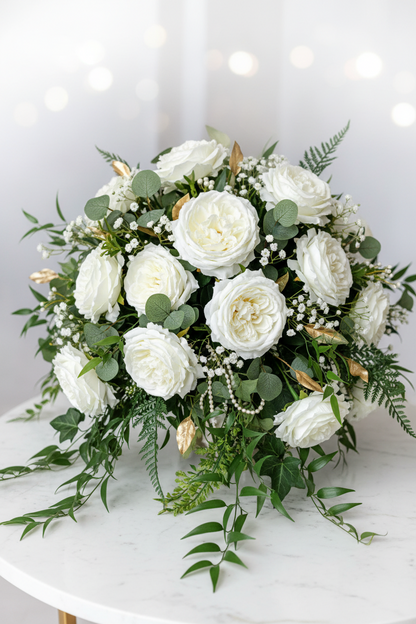 Artificial white silk open roses styled in a lush bridal bouquet with white peonies, gypsophila, green eucalyptus and gold accent buds on a white marble table with soft bokeh background